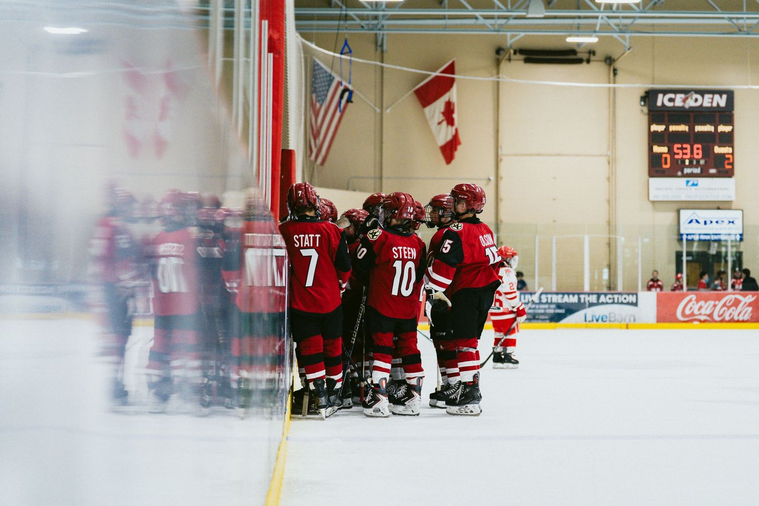 Hockey team in red jerseys huddles together in celebration on indoor rink with Canadian flag visible in background.