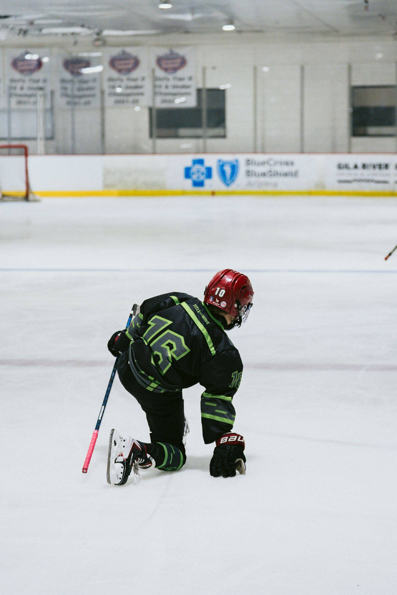 Young hockey player in black uniform kneels on ice rink during a youth hockey game.