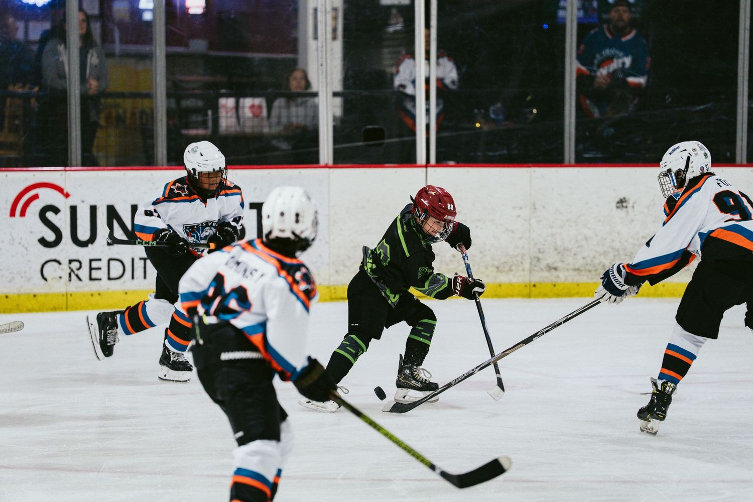 Young hockey players compete on ice, with one player in green controlling the puck against opponents in white jerseys.