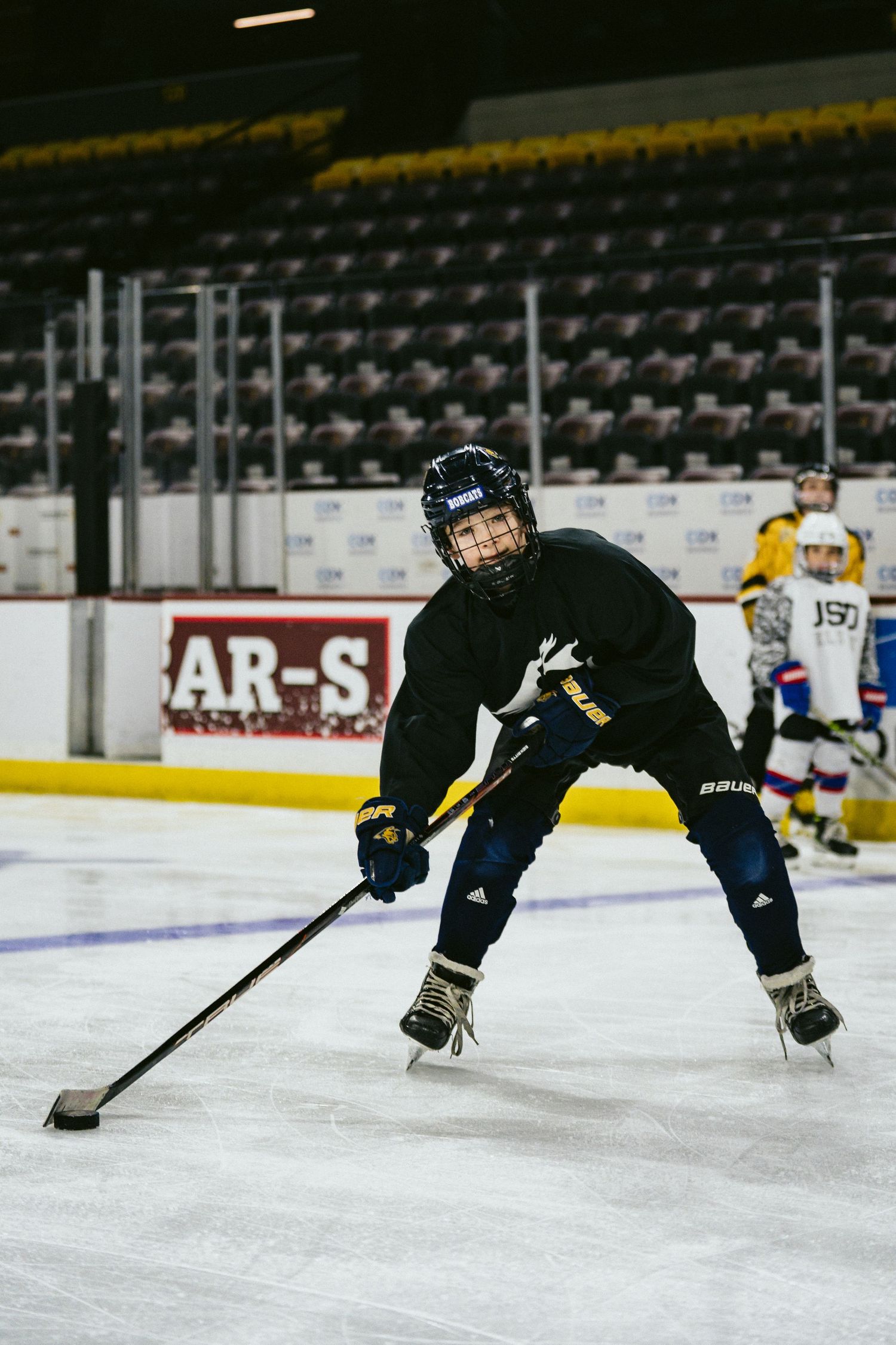Young hockey player in black Bauer gear controlling puck on ice in empty arena.
