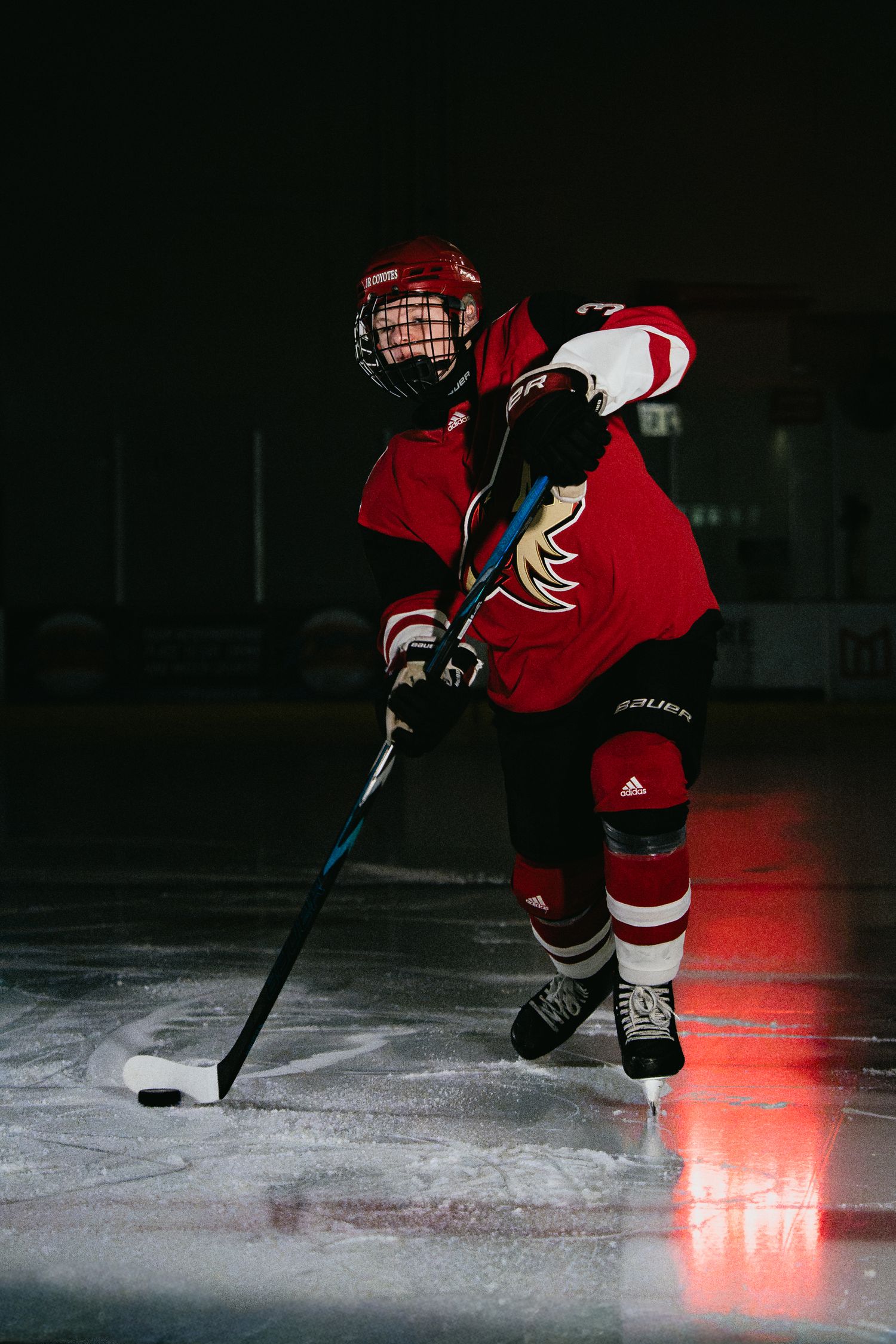 Young hockey player in red uniform skating with stick and puck on ice rink in dramatic lighting.
