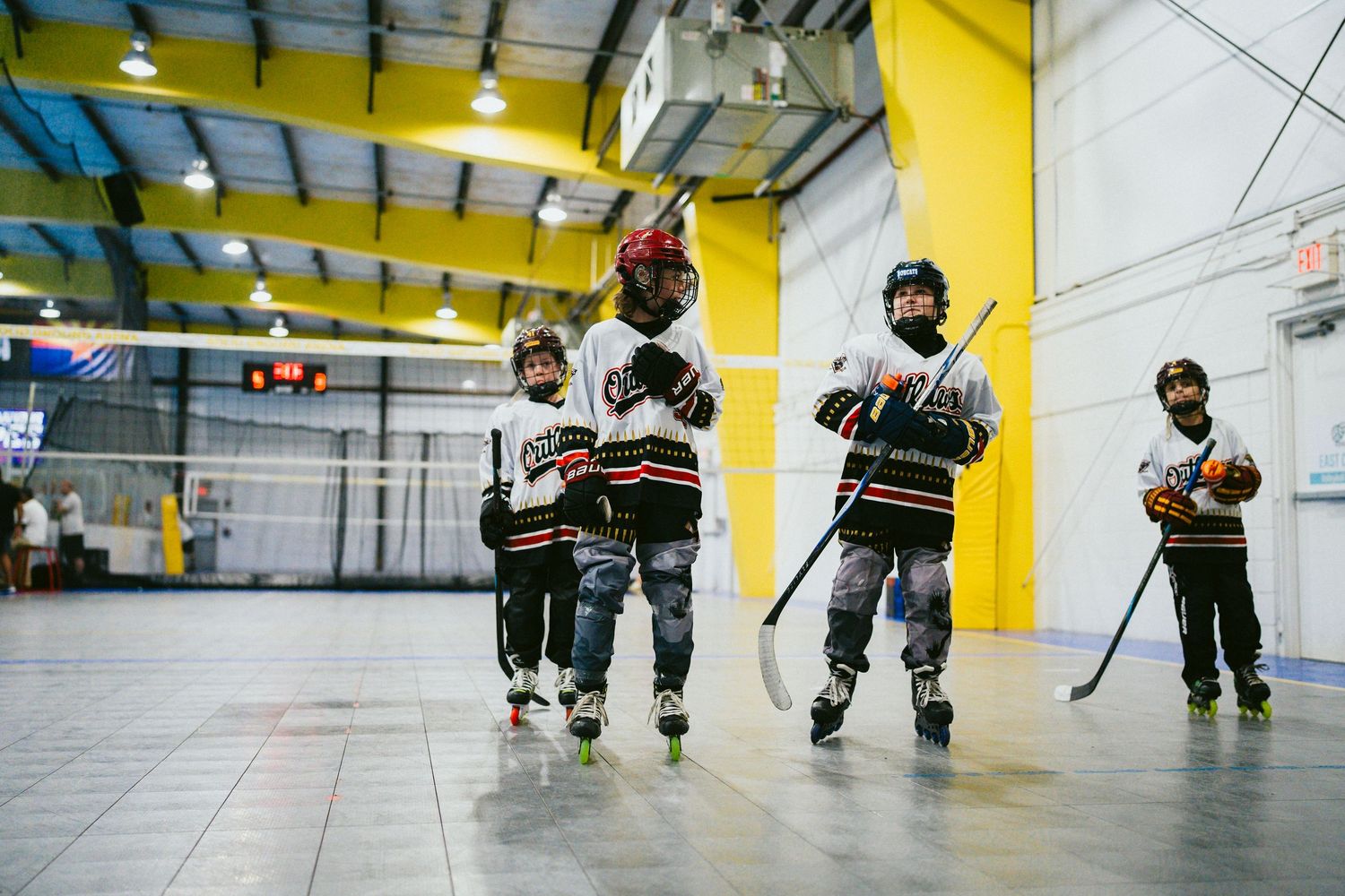 Young children in roller hockey gear practice skating and stick handling in an indoor sports facility.