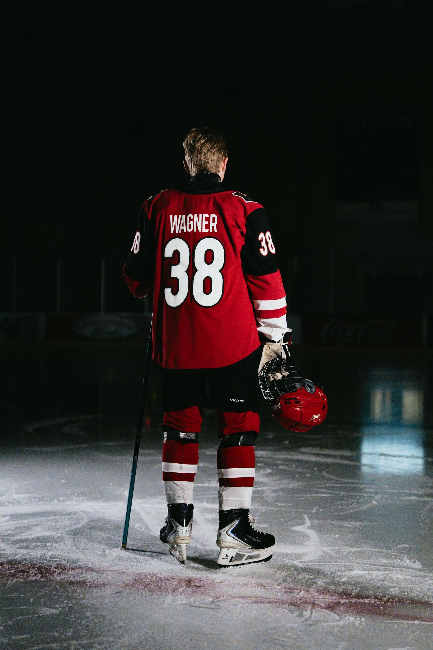 Hockey player Wagner #38 stands on ice rink in red jersey, holding helmet and stick, viewed from behind.