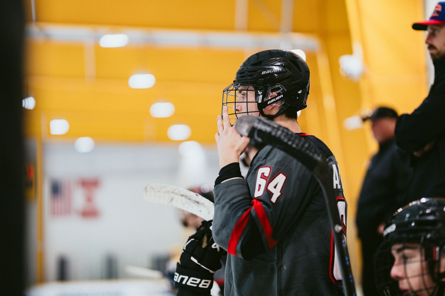 Hockey player number 64 in black and red jersey stands near bench holding stick at rink.