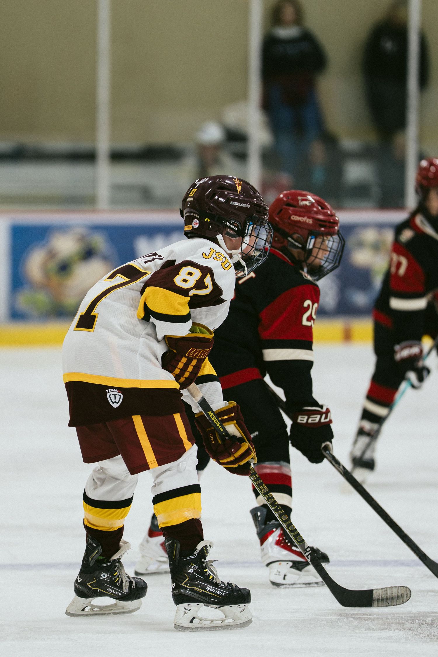 Two youth ice hockey players compete for the puck during a game, wearing full protective gear and skates on an indoor rink.
