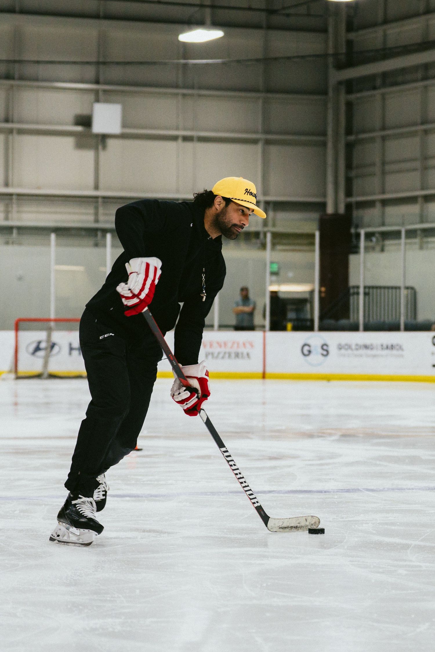 Male hockey player in black outfit and yellow cap handles puck with stick on indoor ice rink during practice.
