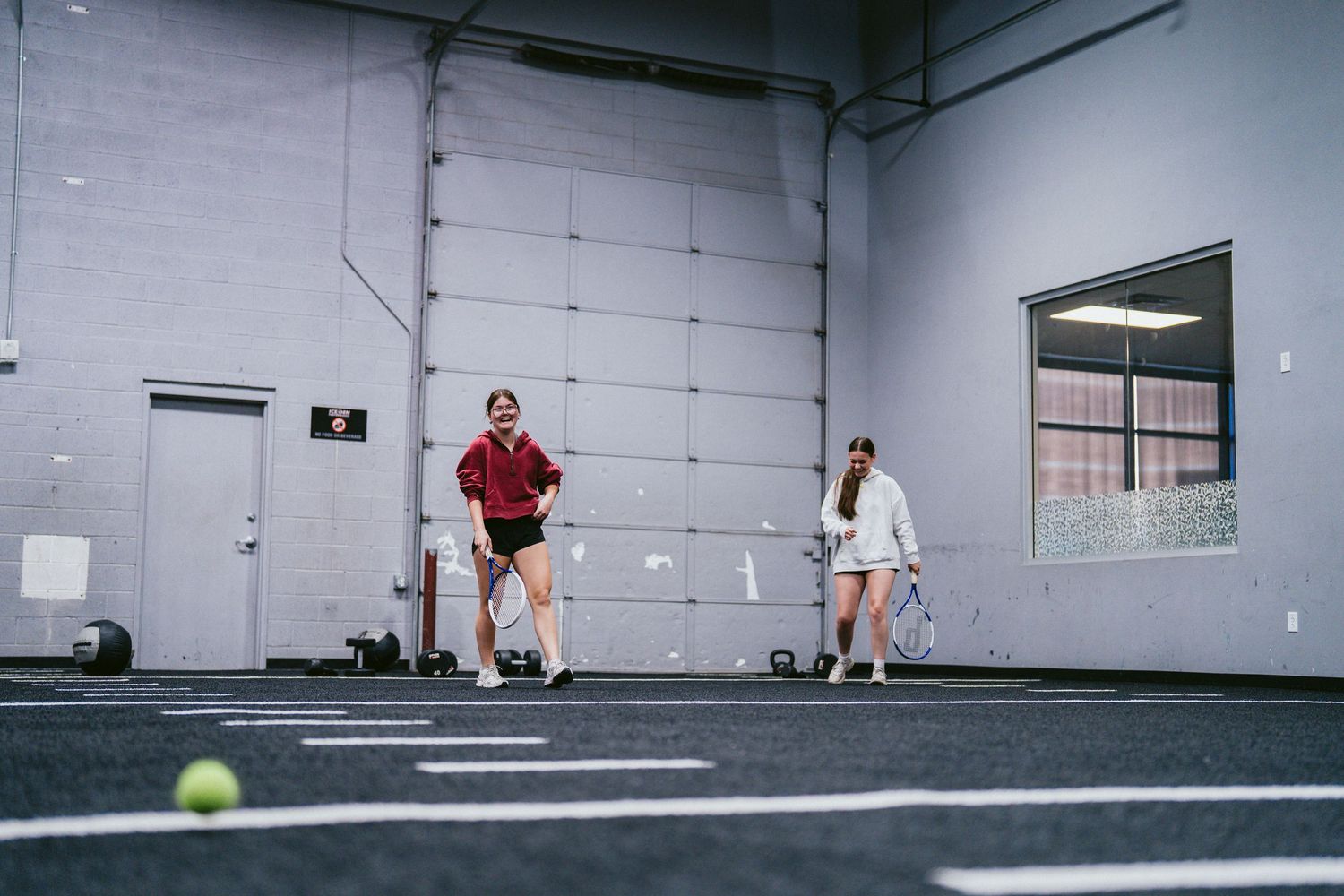 Two women playing tennis indoors in a modern gym facility with turf flooring and a tennis ball in motion.