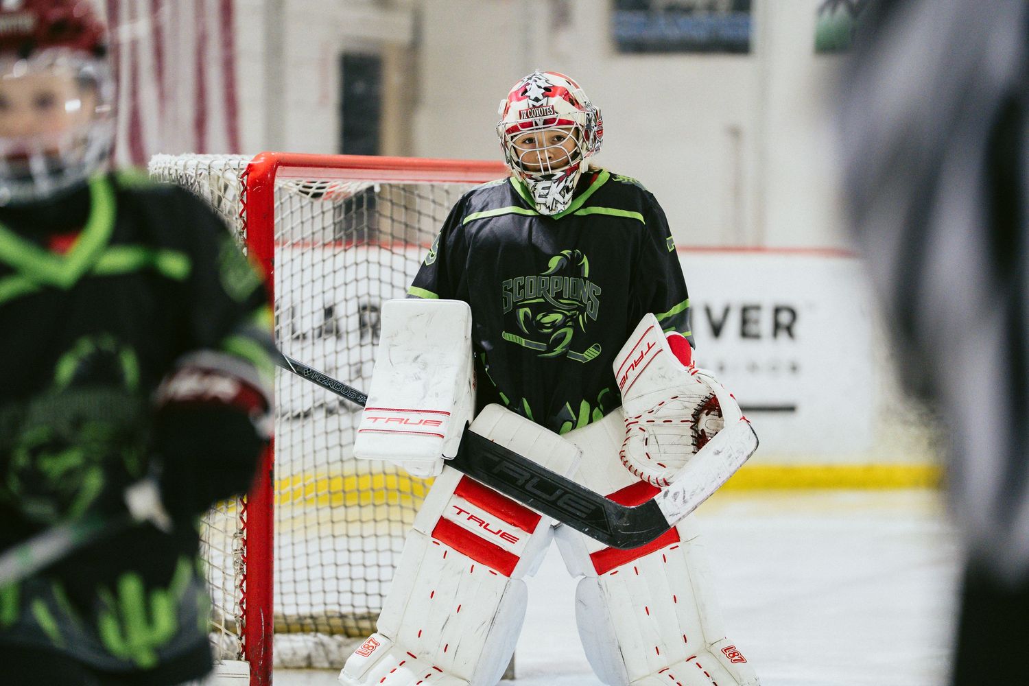 Youth ice hockey goalie in black and green uniform guards the net during a game indoors.