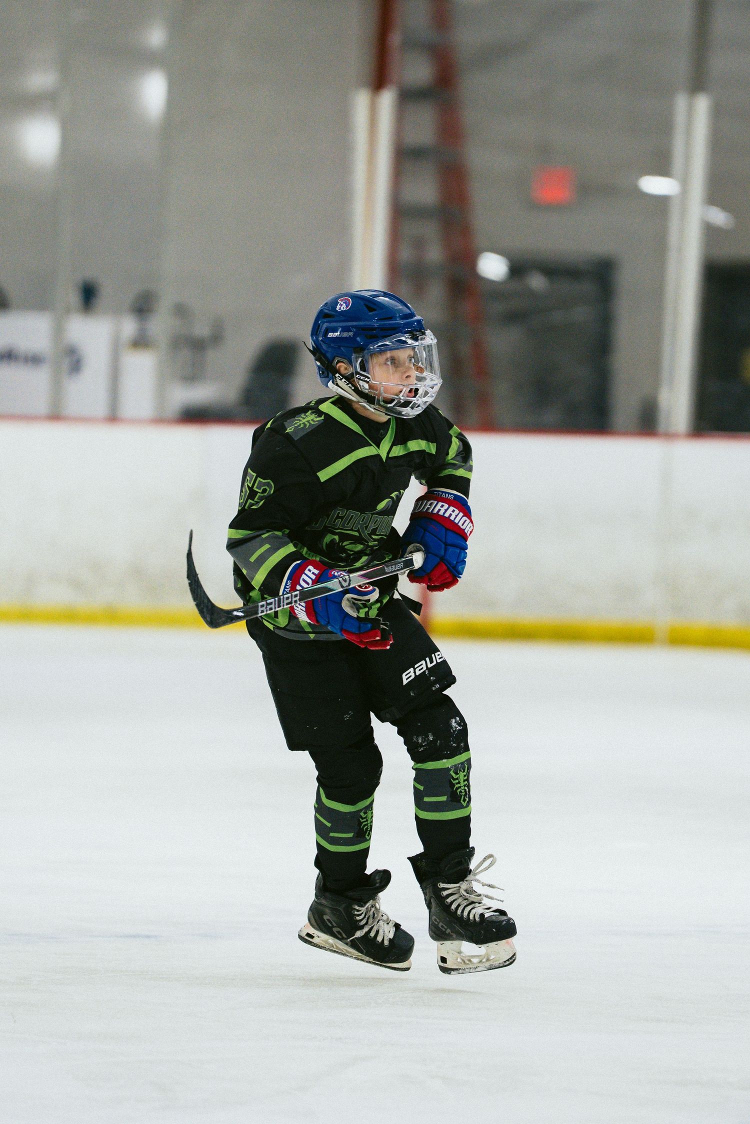 Young hockey player in black and green uniform with blue helmet skating on indoor ice rink.