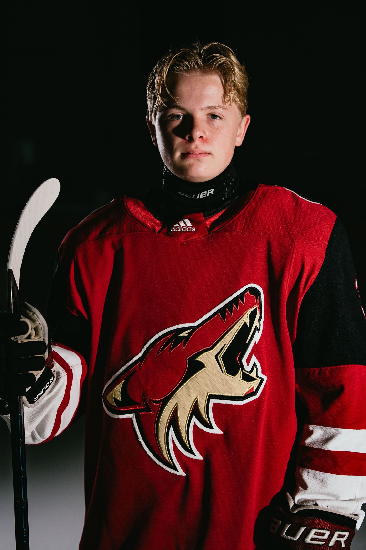 Young hockey player in Arizona Coyotes red jersey holding a stick, posing for an official team portrait.