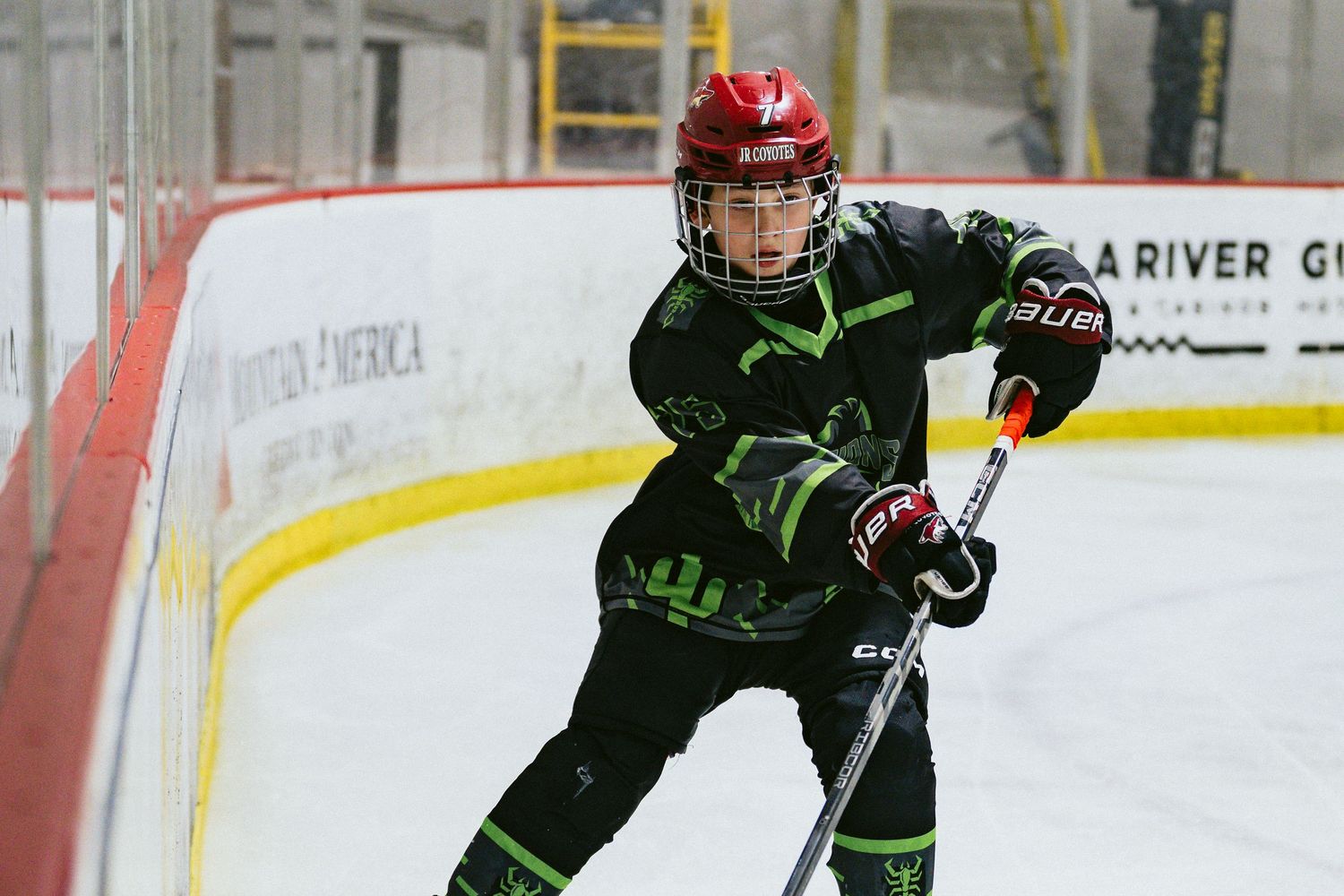 Young hockey player in black and green uniform skates aggressively during an indoor youth hockey game.