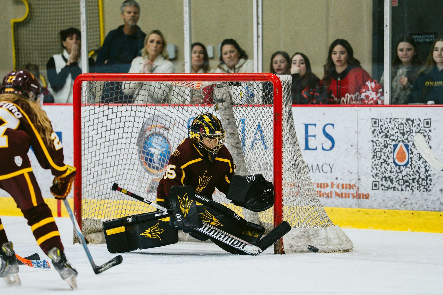 Hockey players compete near the goal with spectators visible through the glass behind them.