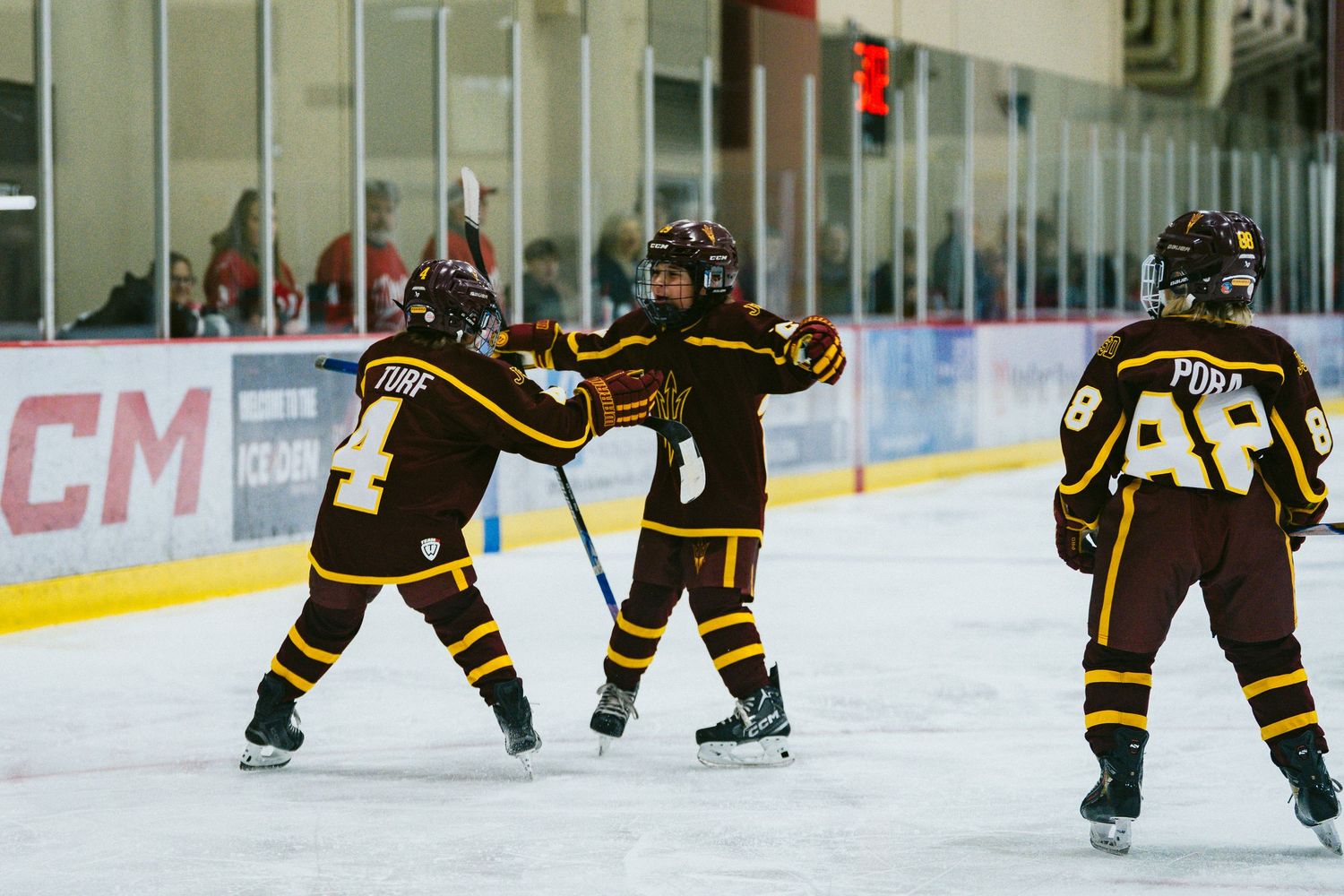 Three youth hockey players in black and gold uniforms practicing drills on an indoor ice rink.