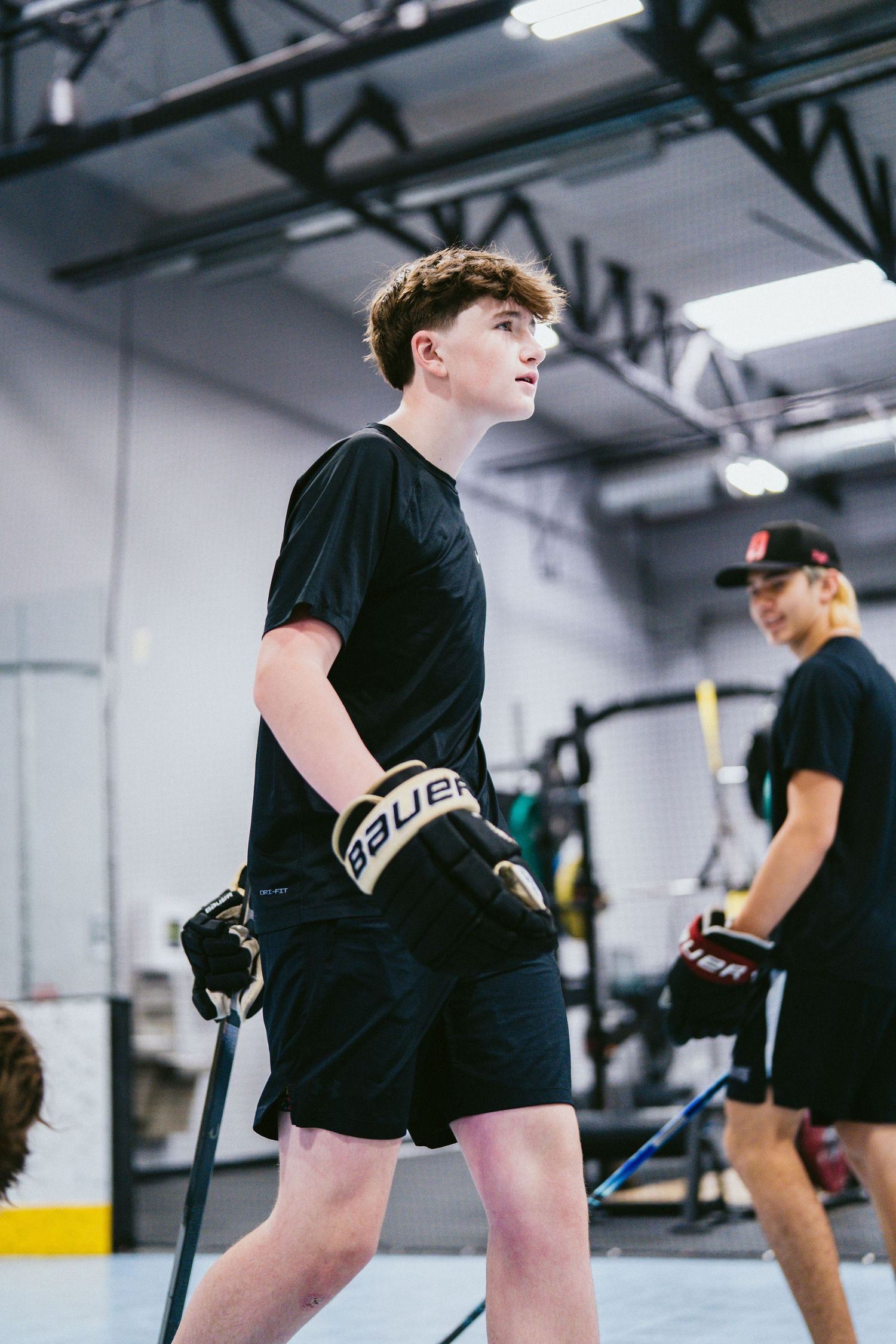 Young hockey player in black outfit wearing Bauer gloves holds a stick during an indoor training session.