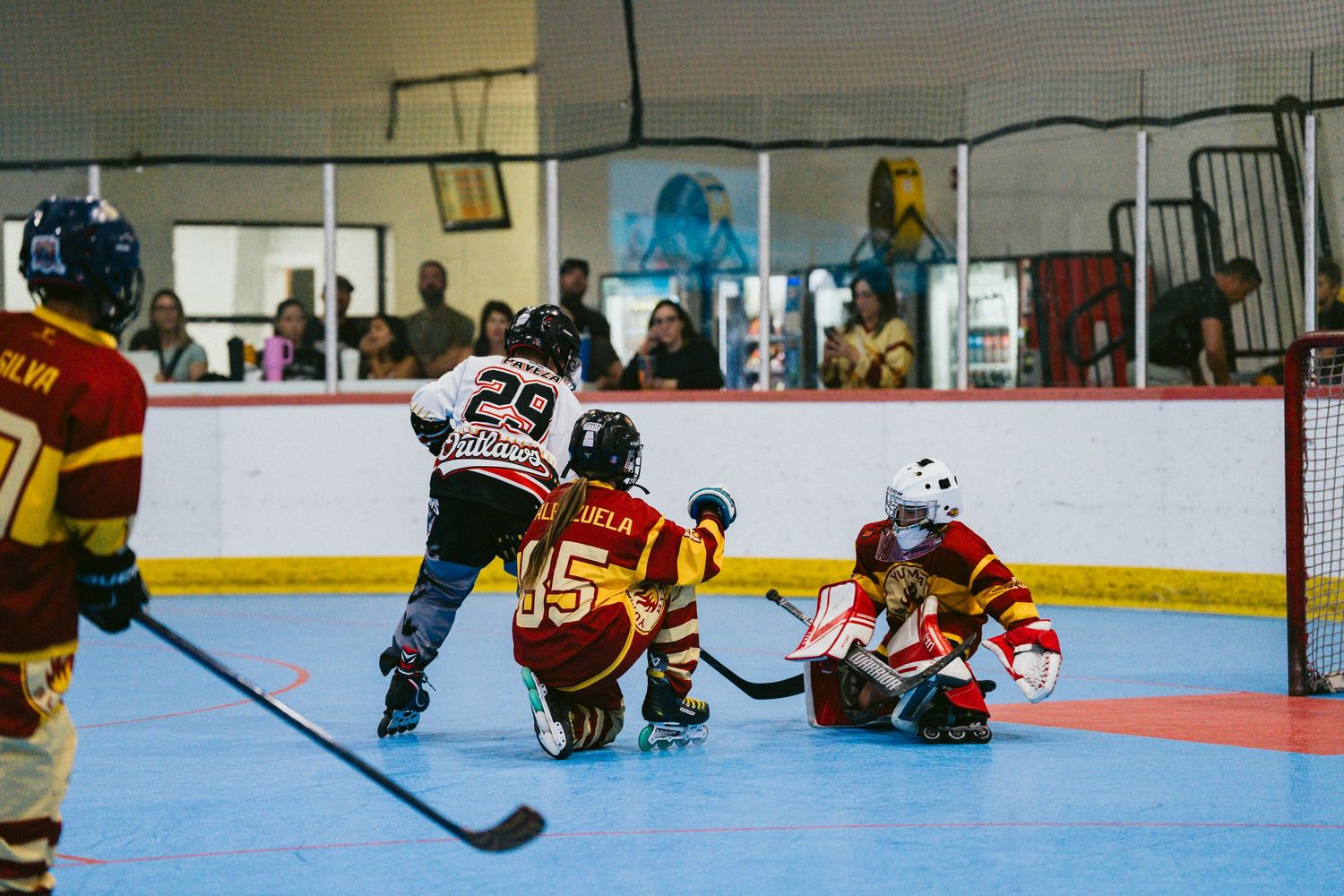 Young roller hockey players battle near the goal as the goalie makes a save on a colorful rink.