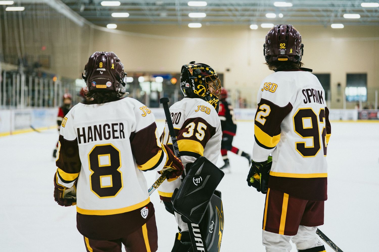 Three youth hockey players including HANGER 8 and SPRIM 92 stand together on the ice rink bench area.
