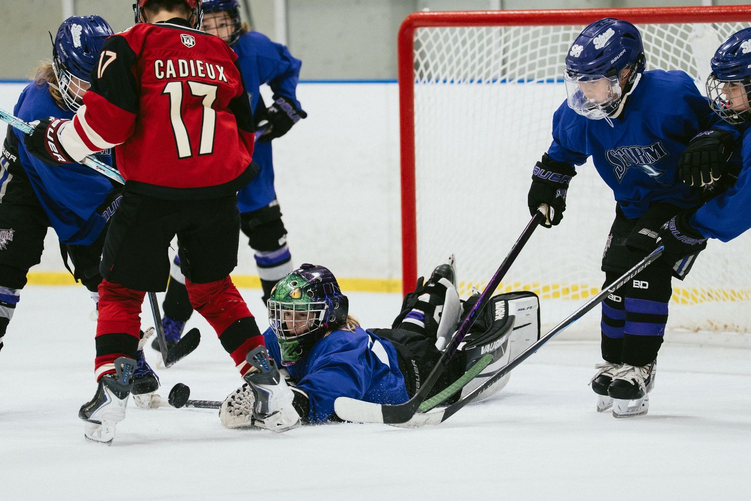 Youth ice hockey players compete intensely, with a goalie diving on the ice to block a puck during a game.