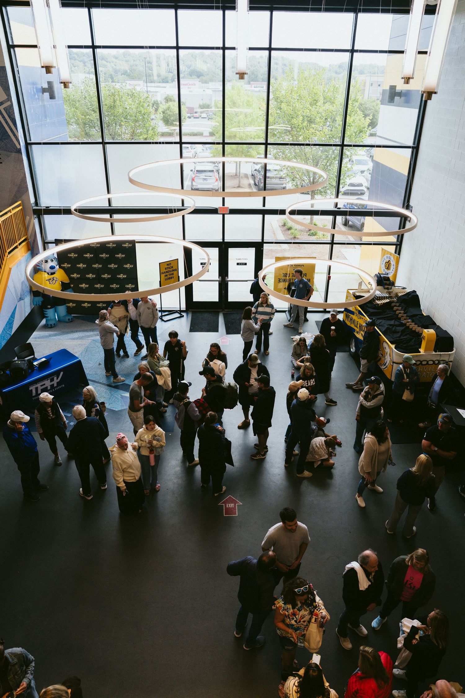Aerial view of a busy indoor venue lobby with crowds of people gathered beneath large windows and circular ceiling fixtures.