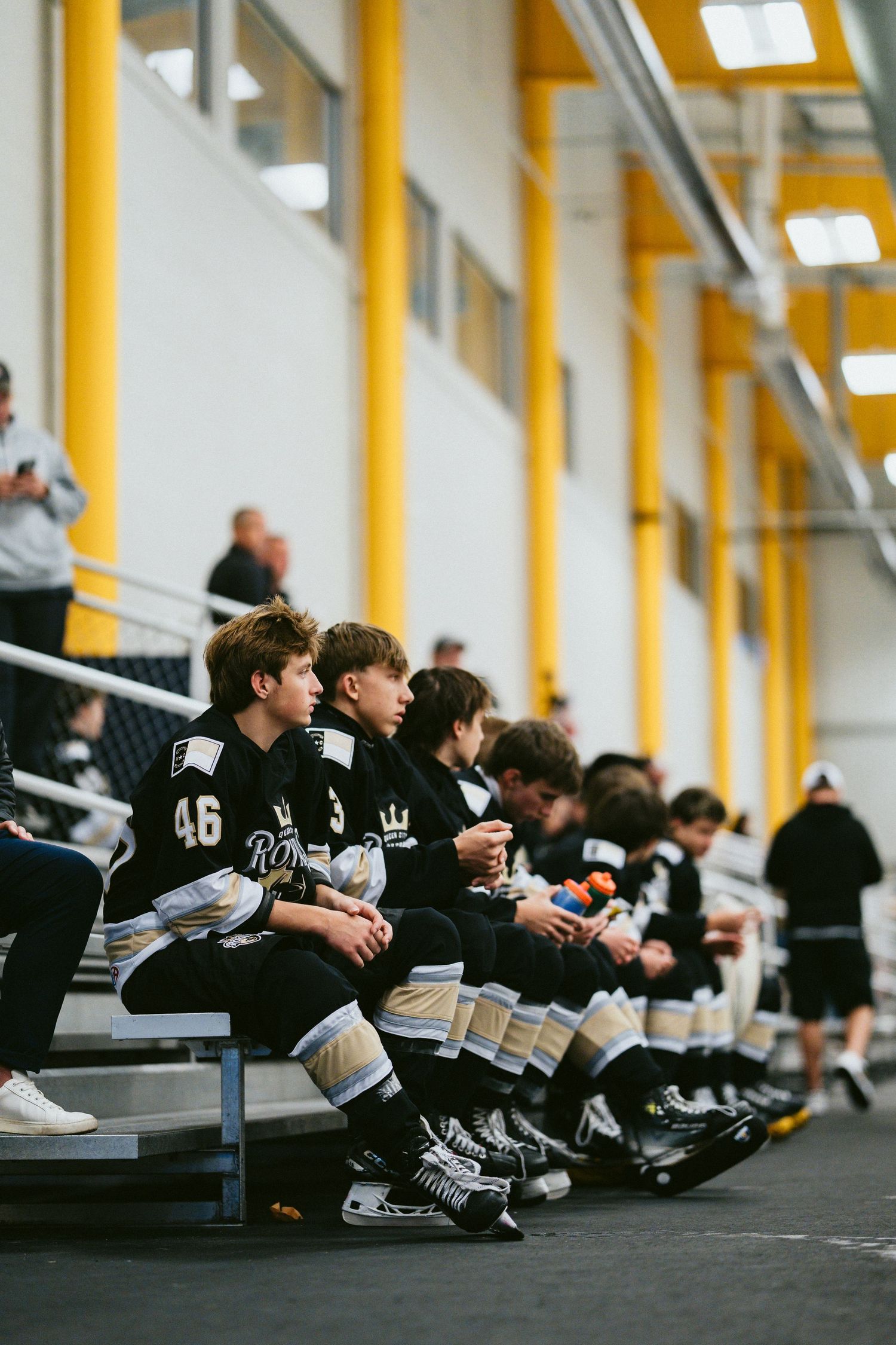 Youth hockey players in black and white uniforms sit on a bench inside a modern hockey facility with yellow accents.