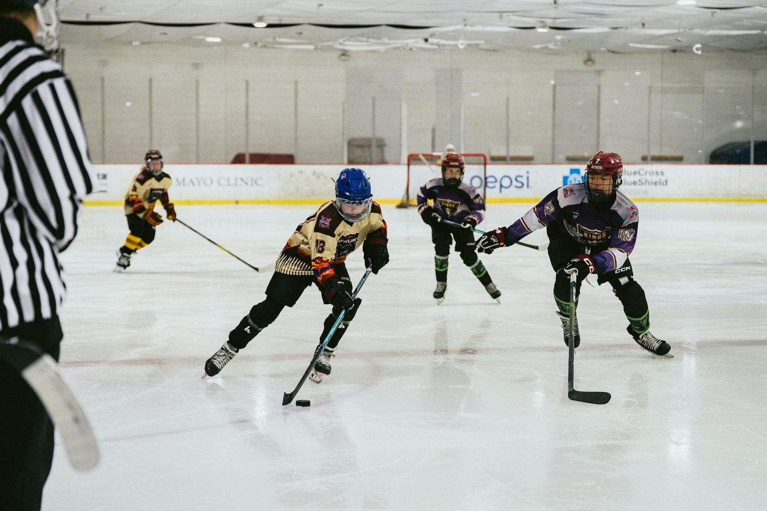 Youth hockey players compete for the puck on indoor ice rink with referee watching nearby.