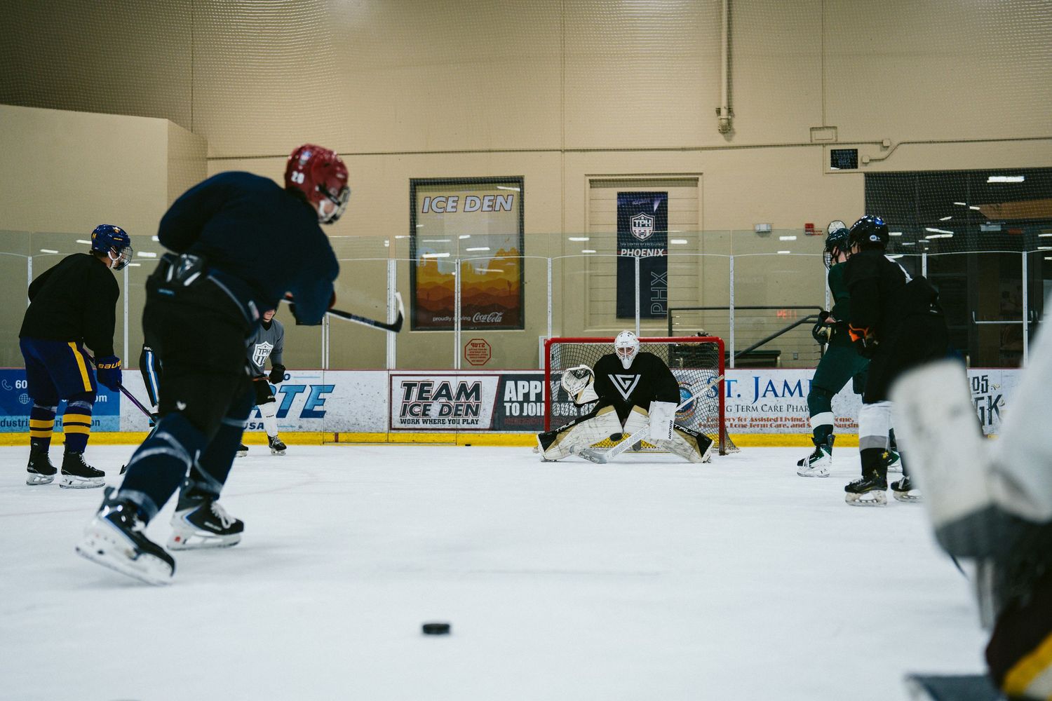 Hockey players practice on an indoor ice rink during a team scrimmage with a goalie defending the net.