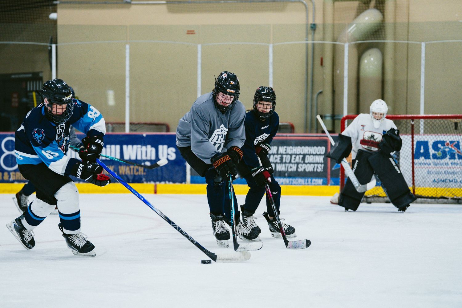 Ice hockey players compete for puck during scrimmage practice at indoor rink with goalie in background.