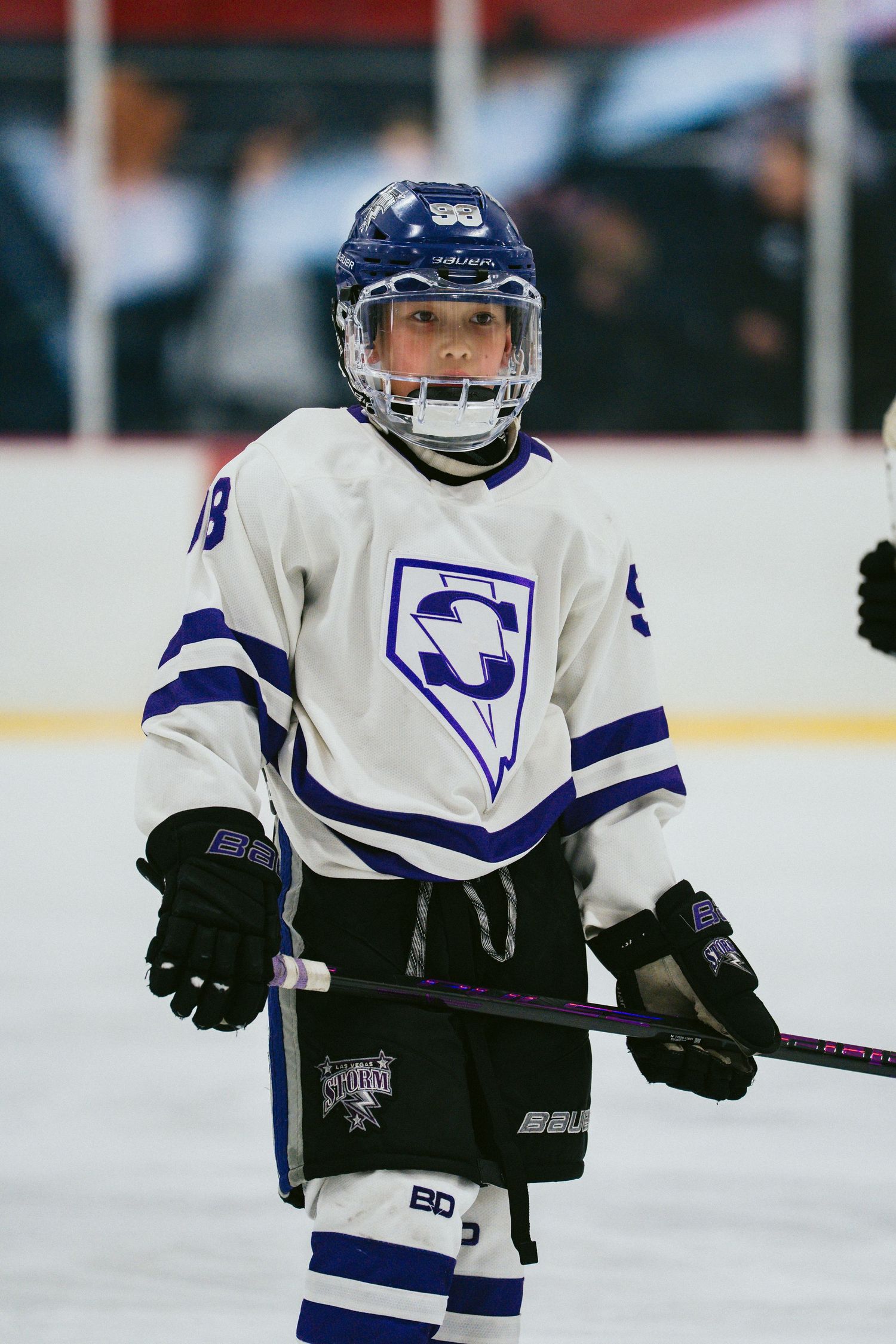 Youth hockey player in white and blue uniform stands ready on the ice.