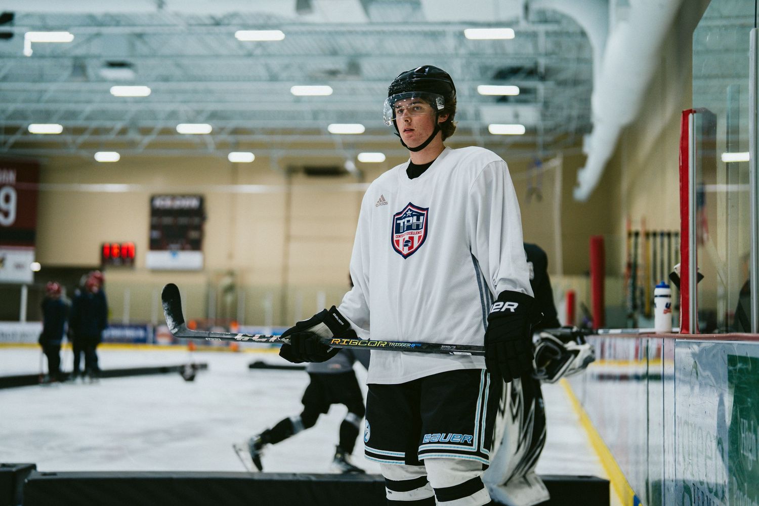 USA Hockey player in white jersey holding a stick on an indoor ice rink during practice.
