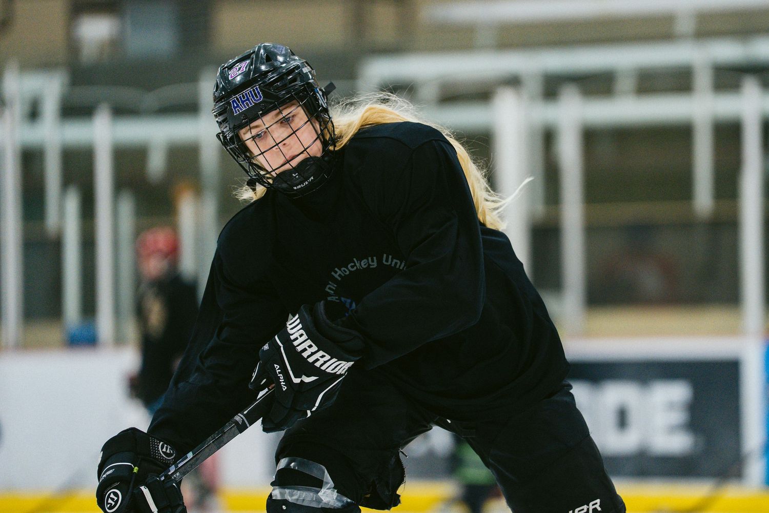 Female ice hockey player in black uniform skating on rink during practice, blonde hair flowing from helmet.