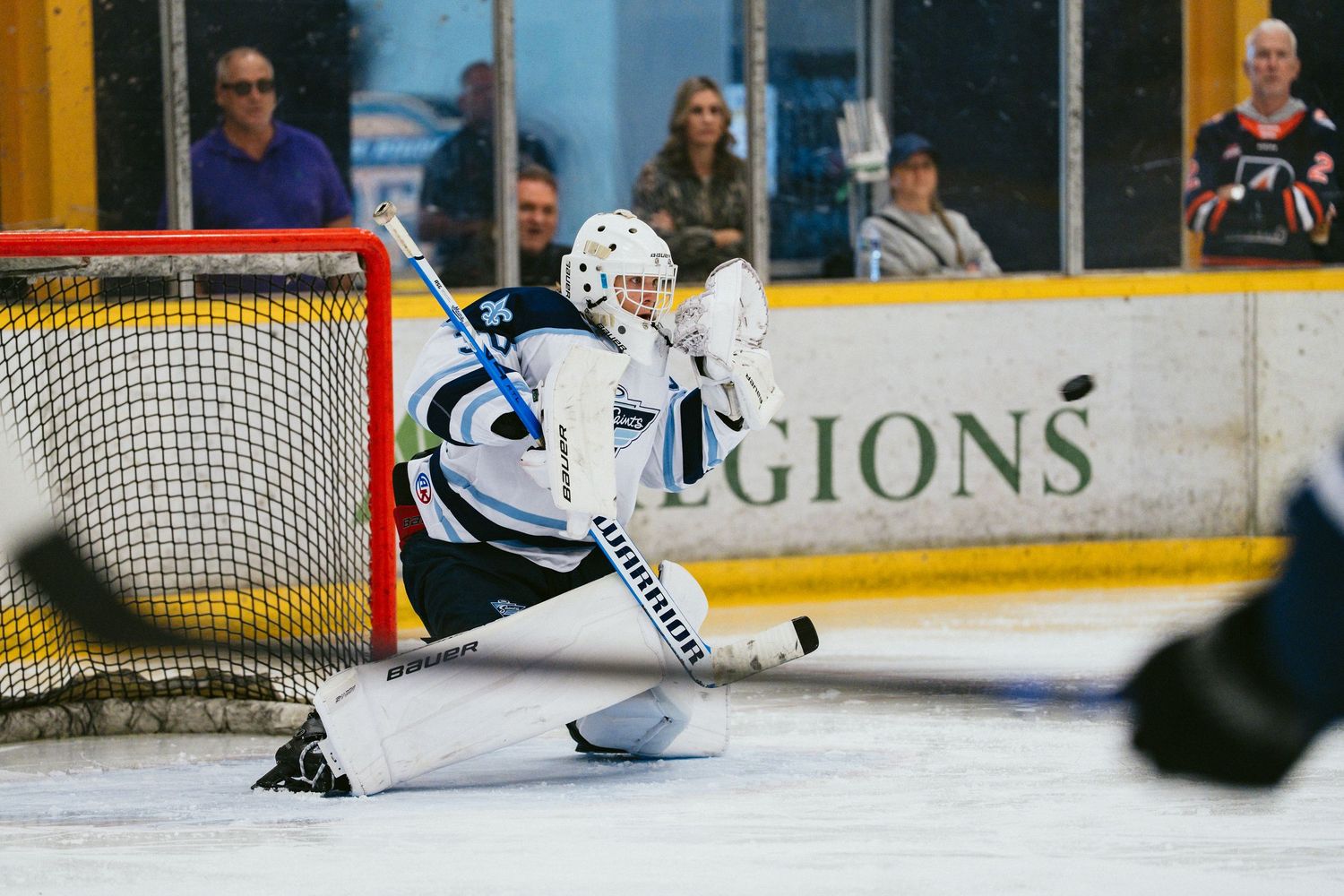 Ice hockey goalie in white and blue gear makes a save attempt during a competitive rink game.