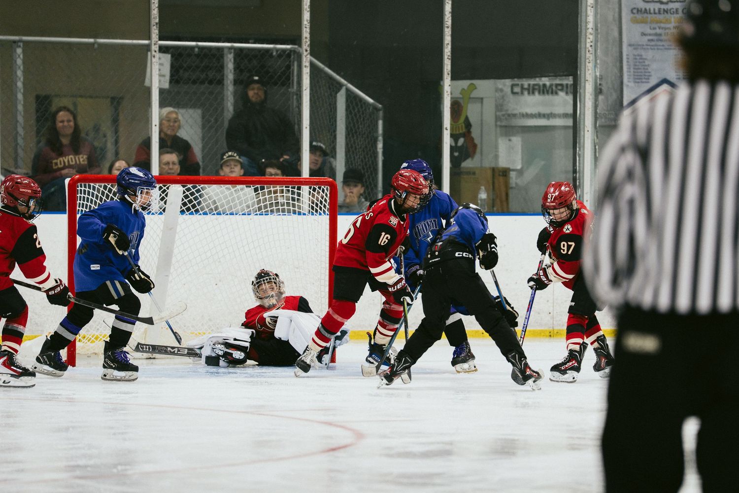 Youth ice hockey players compete near the goal during an intense indoor rink game, with a referee watching closely.