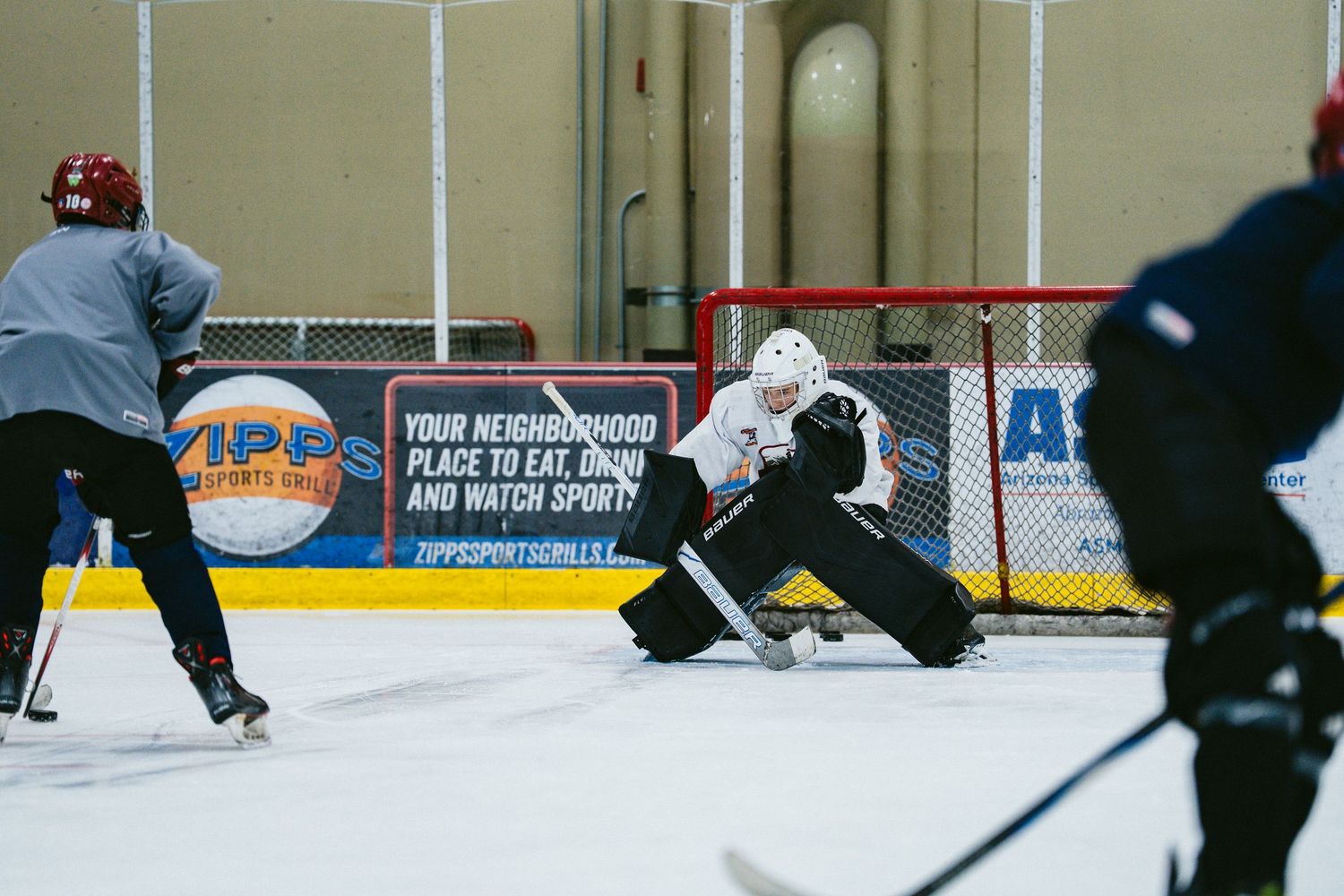 Hockey goalie defends net during indoor ice rink practice session with players approaching.
