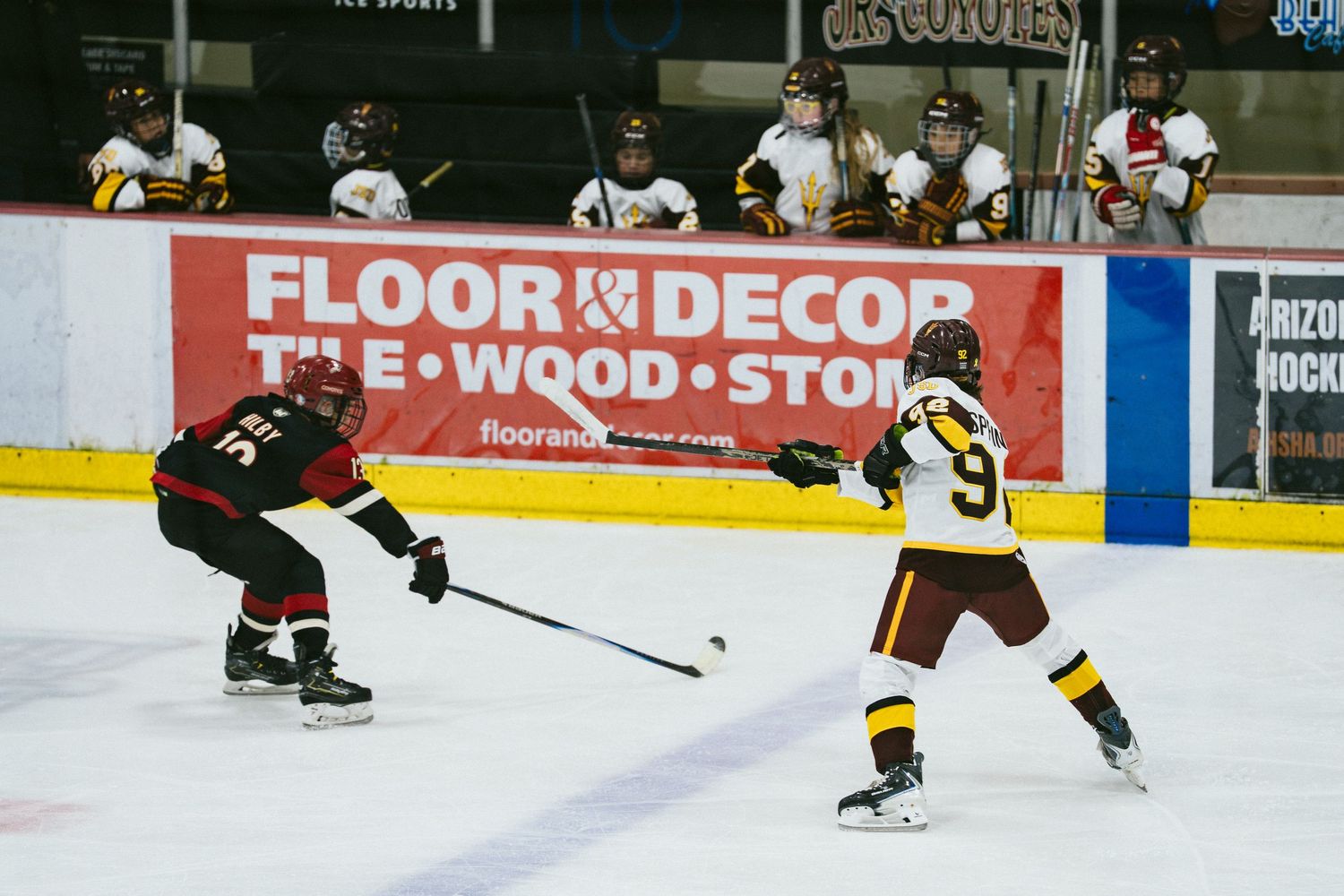 Two ice hockey players compete for the puck during a game near a Floor Decor rink advertisement.