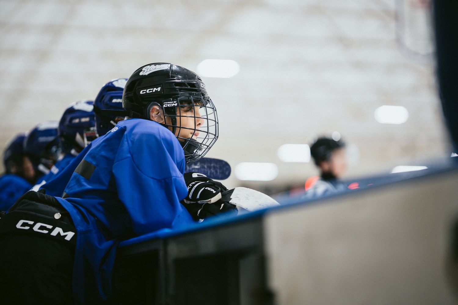 Young hockey player in blue CCM jersey watches game from bench during youth hockey practice.
