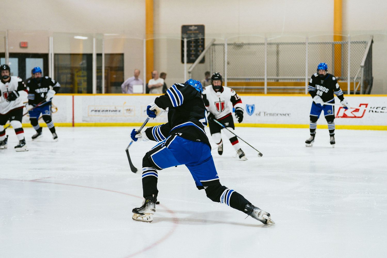 Hockey player in blue uniform takes a powerful shot on ice during an indoor hockey game with teammates nearby.