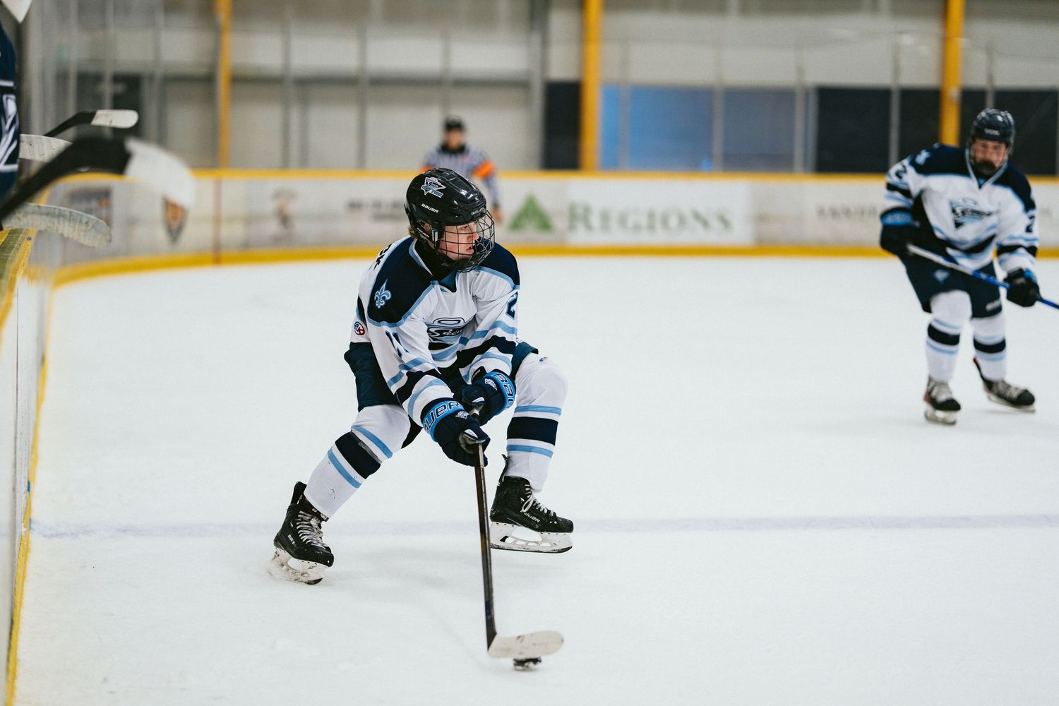 Young hockey player in white uniform controls the puck on ice during a youth hockey game at an indoor rink.