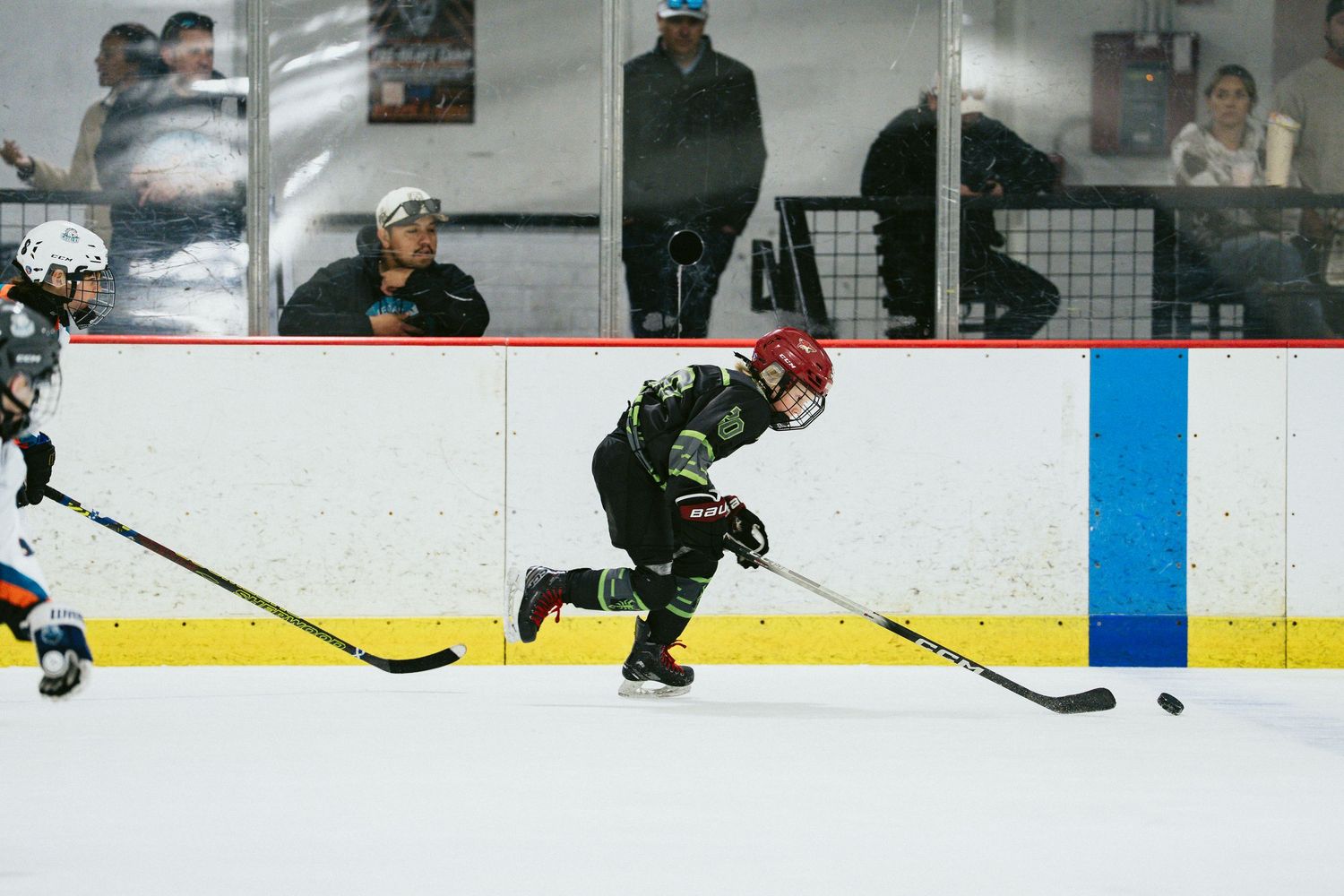 Young hockey player in black green jersey aggressively skates after puck during competitive youth ice hockey game.