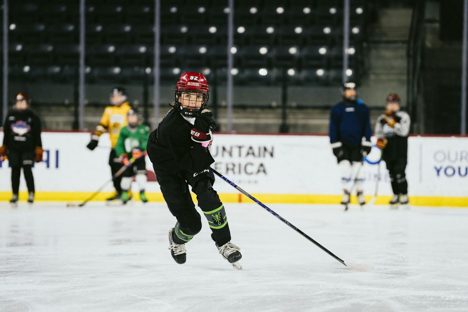 Young youth hockey player skating with stick during practice at indoor ice rink with teammates in background.