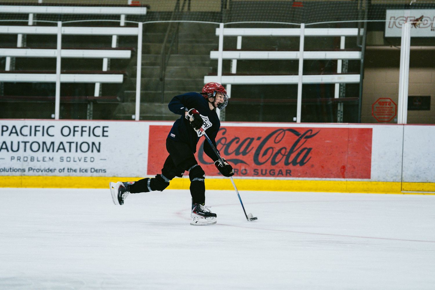 Hockey player in black uniform skates fast across ice rink past Coca-Cola advertisement boards.