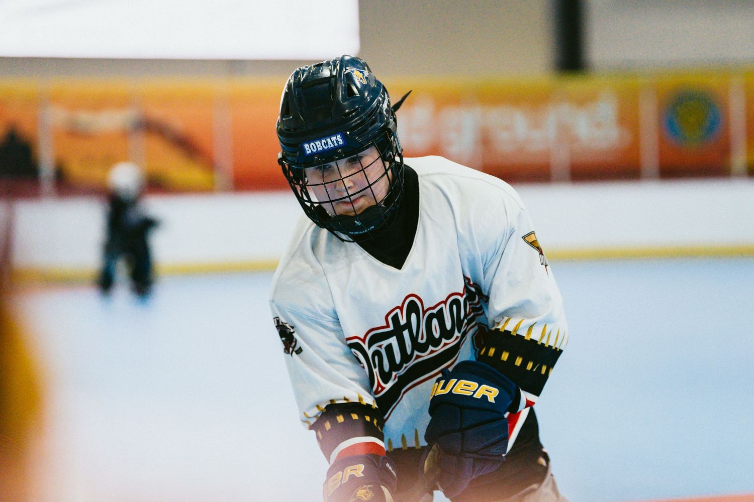 Youth roller hockey player in white Outlaws jersey crouches in ready position during an indoor rink game.