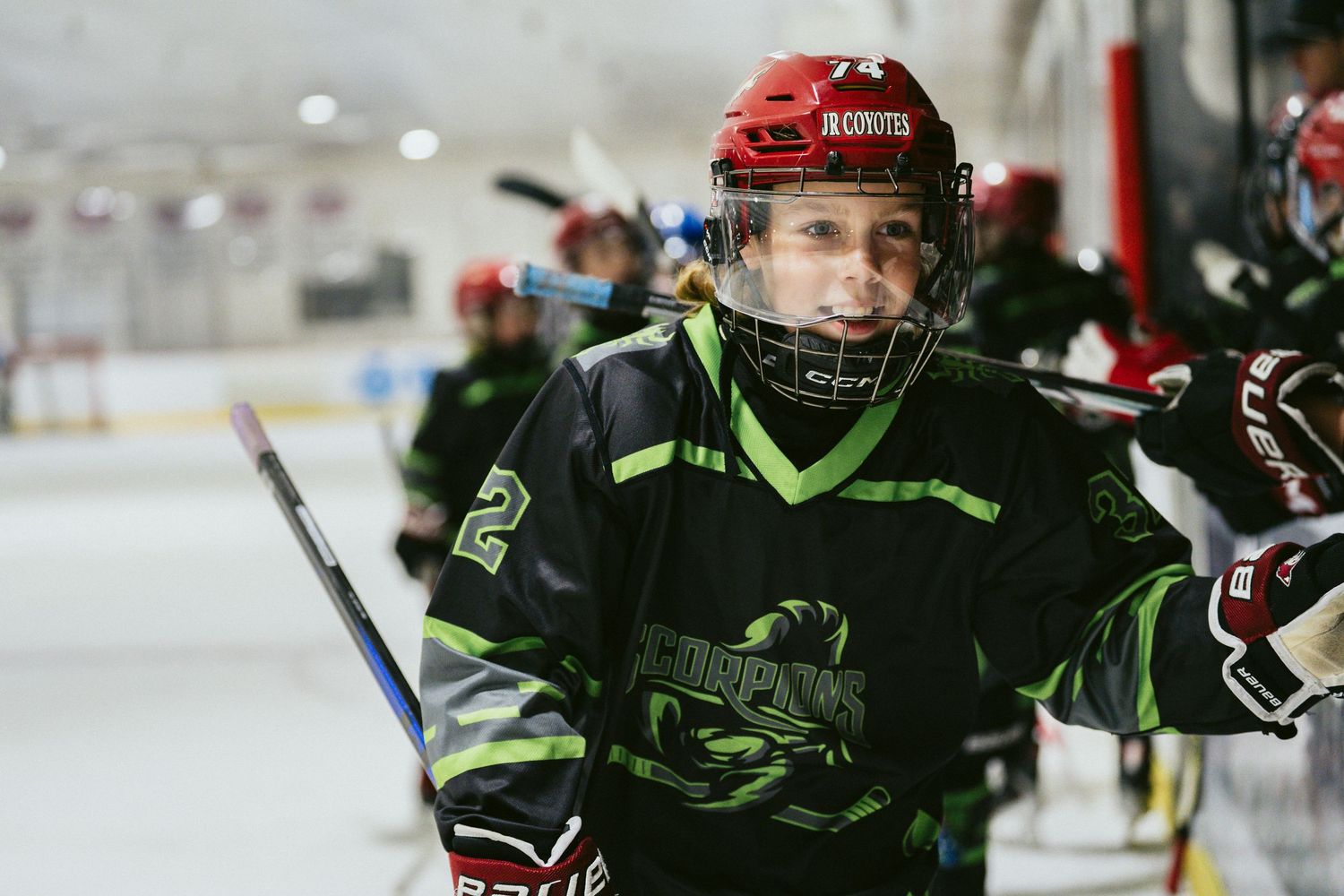 Young hockey player in green Scorpions jersey and red helmet smiling on ice rink during game.