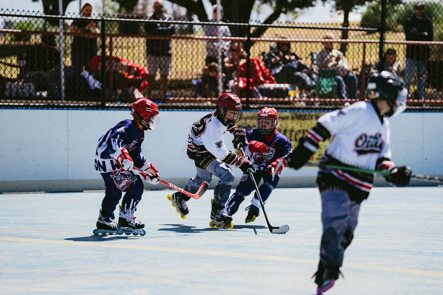 Group of young children in helmets compete in roller hockey game on sunny outdoor rink.