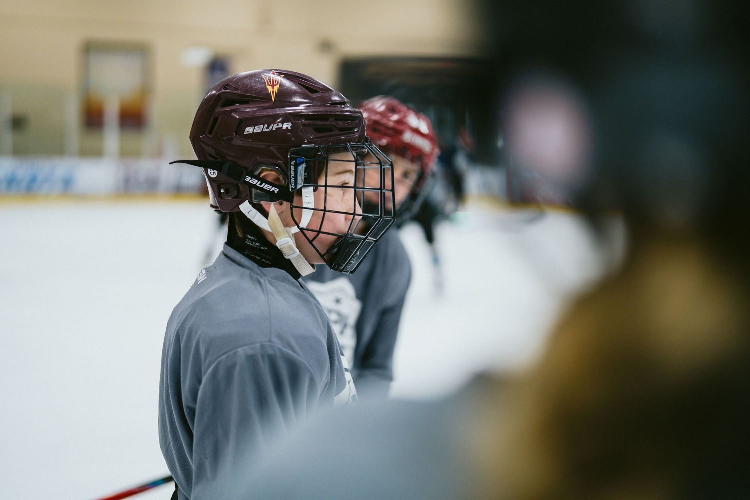 Young ice hockey player in a dark red Bauer helmet listening attentively during a team practice session at an indoor rink.