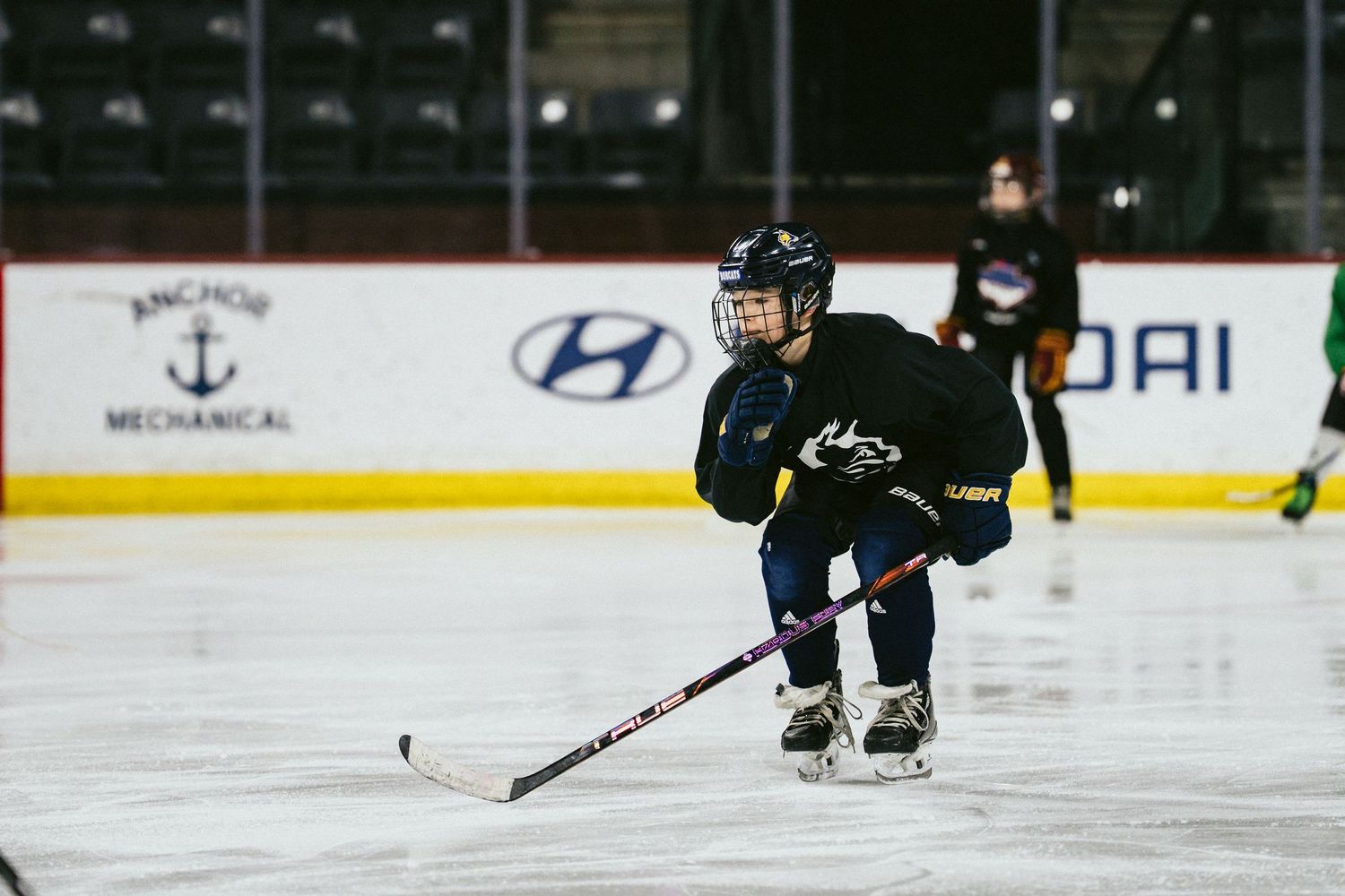 Young child in black jersey skating with a hockey stick during ice hockey practice at an indoor rink.