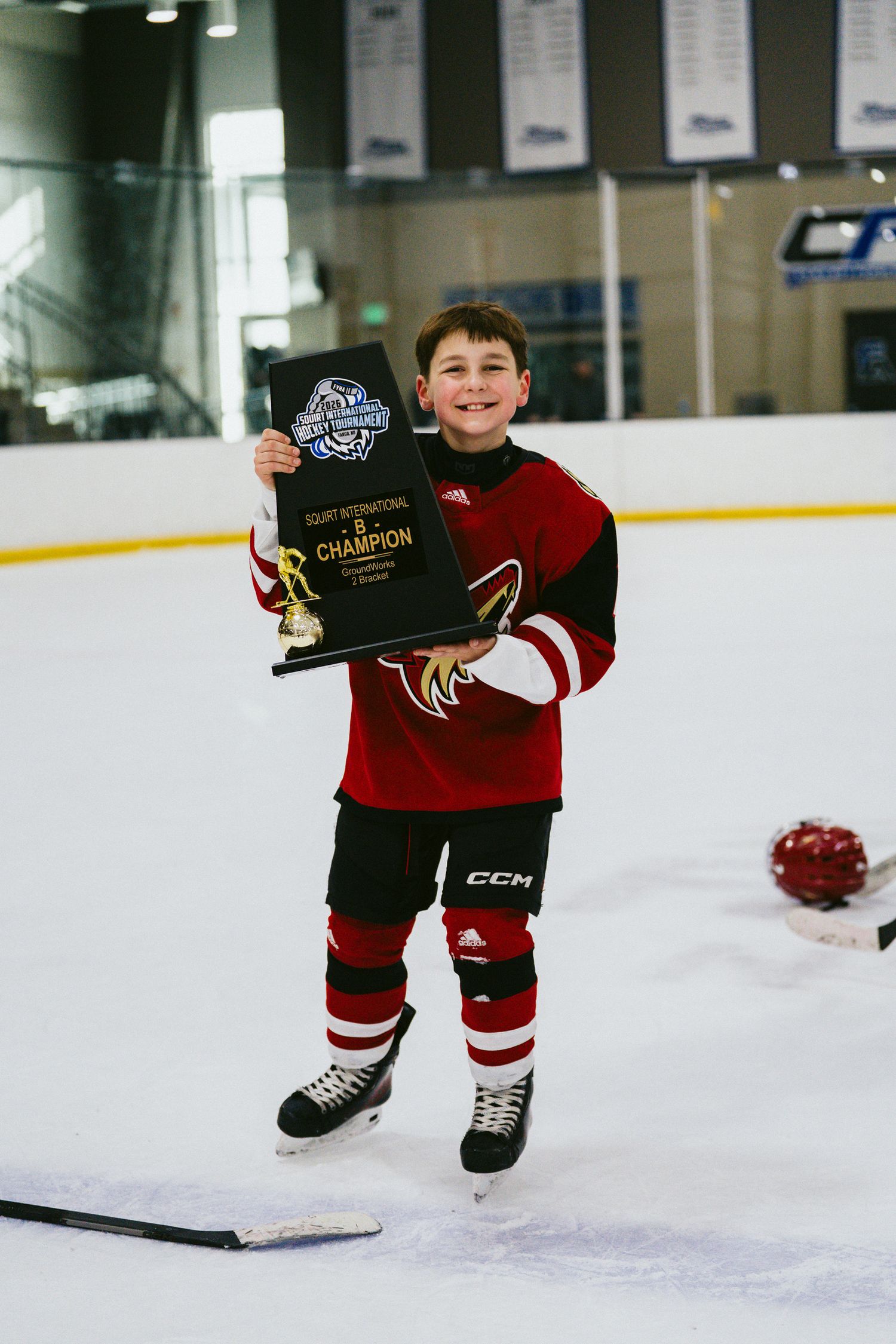 Small athlete in hockey gear displays championship plaque on indoor rink.