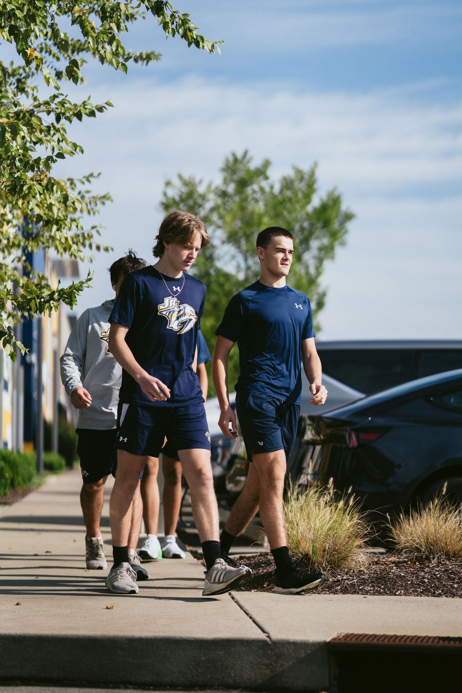 Young hockey players in navy Under Armour gear walk along a sunny outdoor sidewalk near a parking lot.