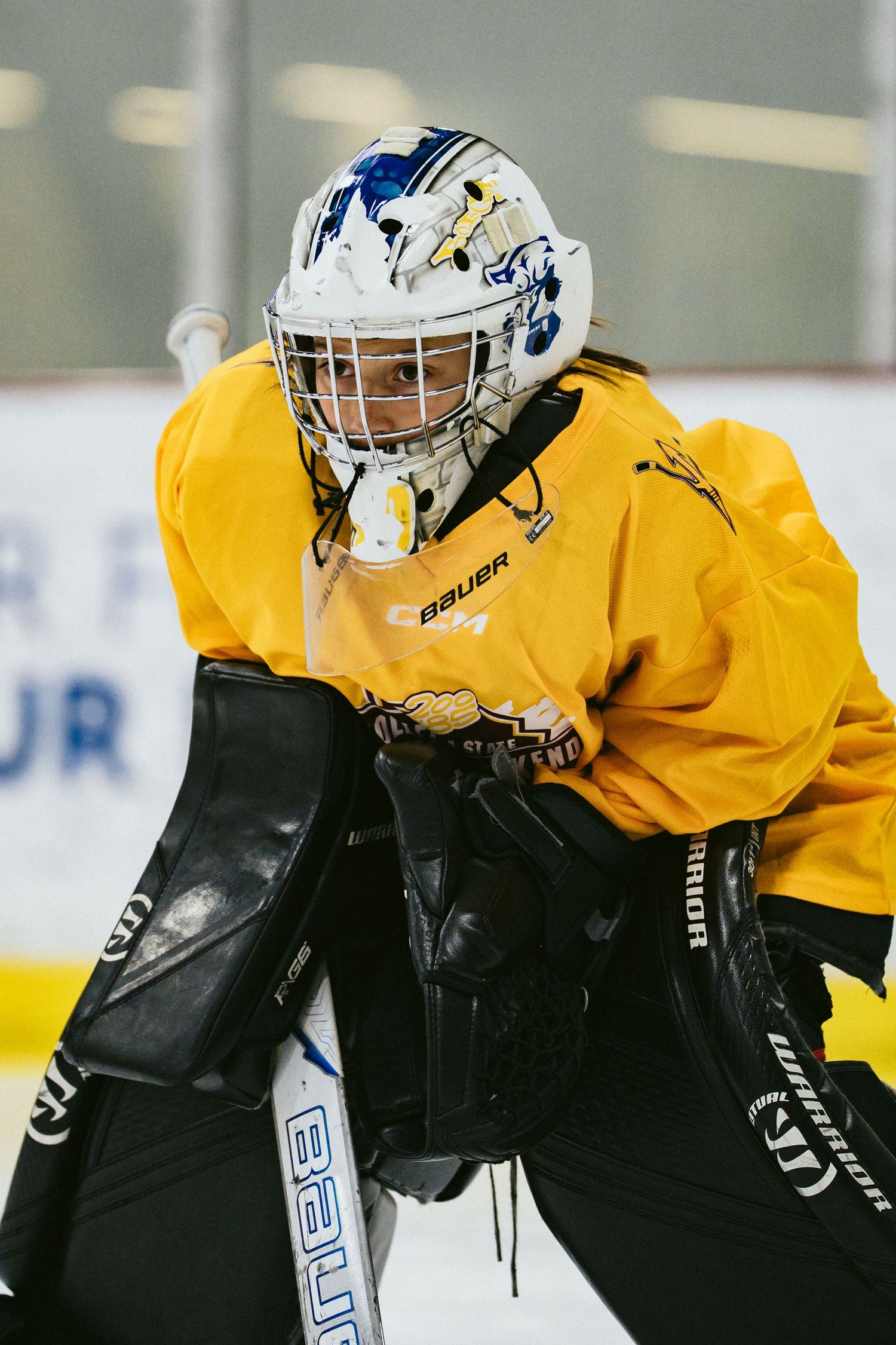 Youth goalie in yellow Bauer gear and decorated helmet ready in stance on ice.