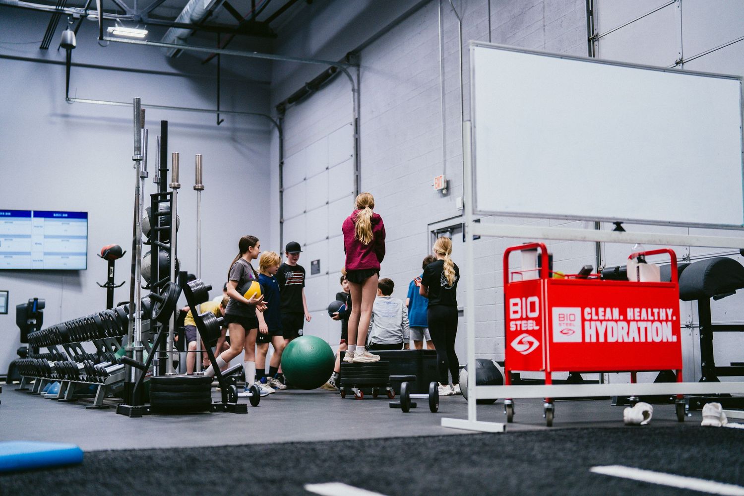 People working out in a modern gym with weightlifting equipment, exercise balls, and hydration station signage.