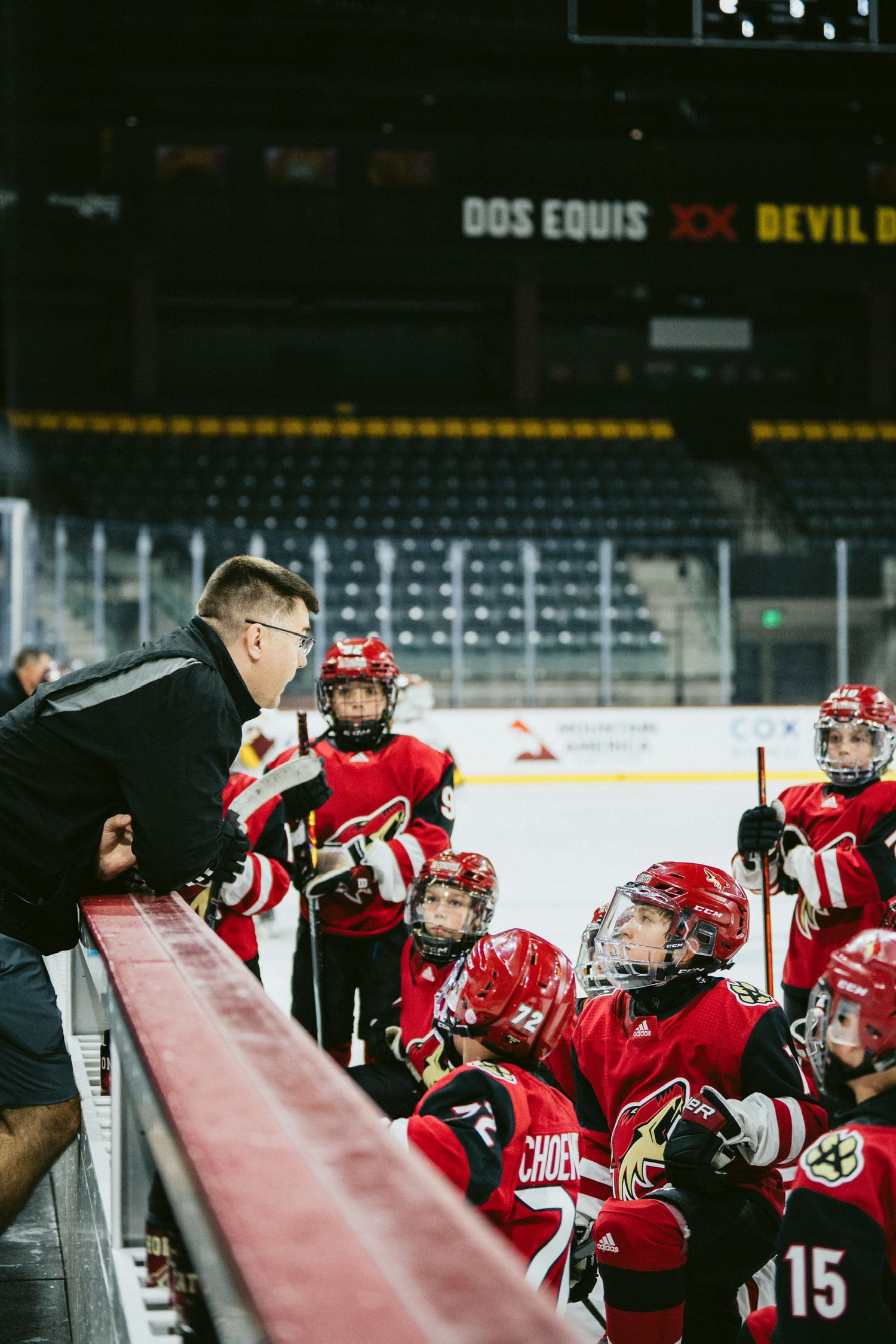 Youth hockey players in red jerseys line up along the boards during a game.
