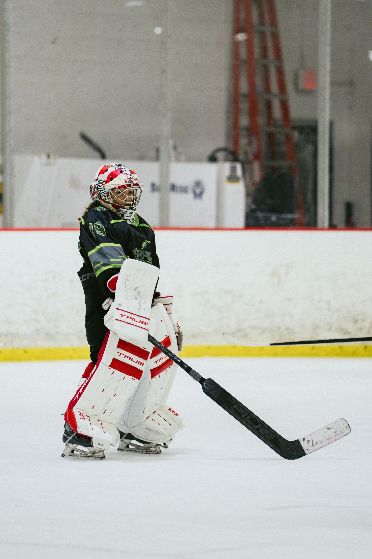 Youth ice hockey goalie in black and white gear stands on ice rink during an indoor hockey game.